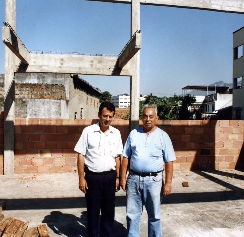 Os Irmãos Montese e Amorim, de camisa clara, posam de pé sobre a laje, diante de uma parede de tijolo furado ainda sem reboco. Atrás, um pórtico de concreto, telhados do bairro e os morros ao longe.