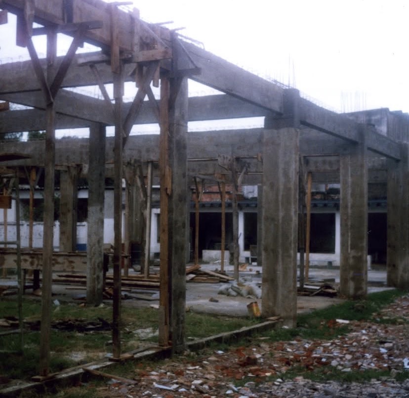 Pórticos de concreto sustentados por escoras de madeira roliça, vistos do pátio interno da obra. No chão, cacos de tijolo e restos de tábua.