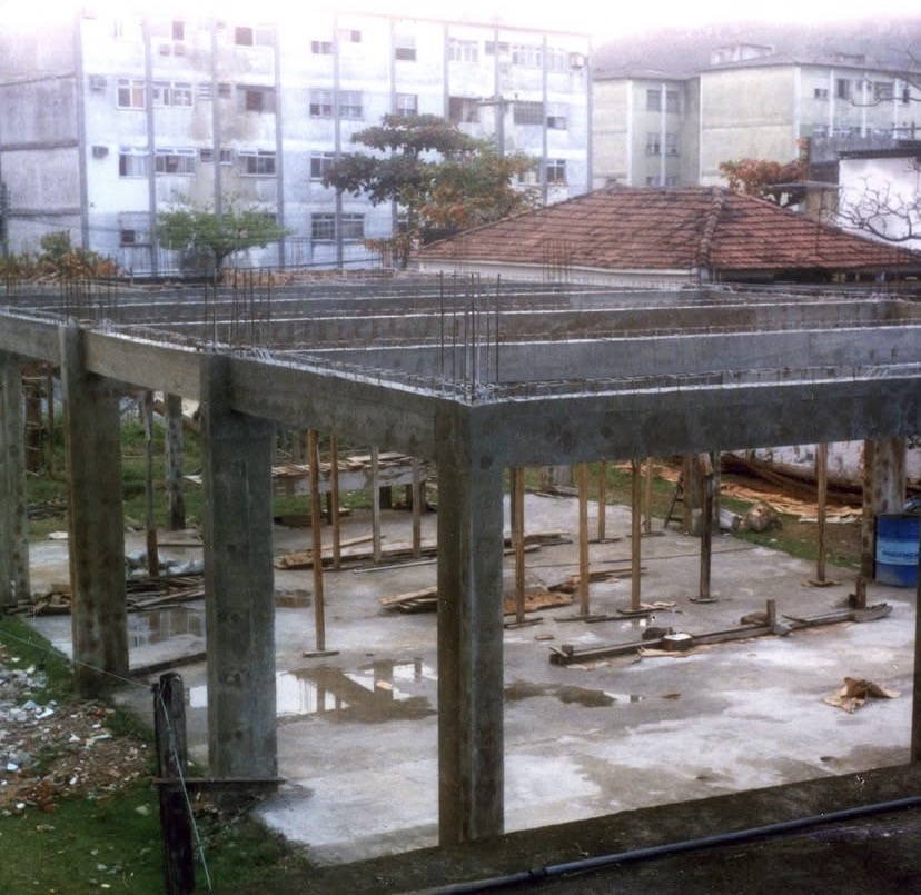 Laje armada apoiada em pilares de concreto, com barras de ferro de espera apontando para cima. Ao fundo, prédios residenciais e um telhado de telha cerâmica.